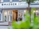 Gadebillede af Madsen Optik, en brillebutik med store glasvinduer, udstillingshylder med briller og en grøn busk i forgrunden.