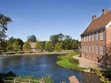 En stor murstensbygning står ved siden af en rolig dam, omgivet af grønne træer og græs under en klar blå himmel. Et mindre rødt hus ses i baggrunden.