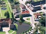 Luftfoto af en lille by med bygninger med røde tage, en dam, velplejede haver og krydsende veje omgivet af grønne områder.