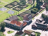 Luftfoto af en lille by med bygninger med røde tage, grønne haver, geometriske hække og en dam med blomsterformede elementer. Gader og træer omgiver området.
