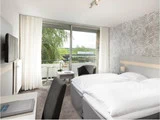 Moderne hotelværelse med to enkeltsenge, mønstret accentvæg, skrivebord, stole, tv og stort vindue med gennemsigtige gardiner med udsigt til en balkon og grønne områder udenfor.