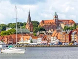 Farverige bygninger og en kirke med et højt spir ligger langs havnefronten i en europæisk by med en sejlbåd, der flyder på vandet i forgrunden.