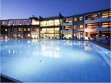 En stor, oplyst udendørs swimmingpool foran en moderne hotelbygning i flere etager i skumringen.