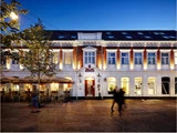 Et veloplyst hotel i to etager med udeservering, store vinduer og mennesker, der går forbi på en brostensbelagt gade i skumringen.