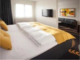 Et moderne hotelværelse med en stor seng med gule puder og tæppe, et væghængt tv, abstrakt kunst, en sofa og et mønstret tæppe. Naturligt lys kommer ind gennem et vindue.