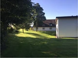 En græsklædt gård med sollys og skygger, omkranset af træer, ved siden af to hvide bygninger med brune tage under en klar blå himmel.