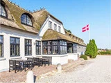 En hvid bygning med stråtag og sortindrammede vinduer ligger ved siden af udendørs borde; et dansk flag vajer i nærheden under en klar blå himmel.