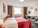Et hyggeligt hotelværelse med en dobbeltseng med rødt og hvidt sengetøj, to grå stole, et tv, et skrivebord, en lampe og et vindue med stribede gardiner.