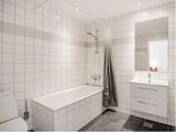 Moderne badeværelse med hvide fliser, badekar, gråt bruseforhæng, toilet, væghængt spejl og hvidt vaskeskab med skuffer.