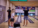 Fire unge mennesker står ved en bowlingbane; den ene med armene i vejret for at fejre, mens de andre holder øje med banerne.