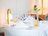 Et pænt dækket restaurantbord med hvid dug, flere glas, sølvtøj, en serviet, en lampe og en enkelt rød rose i en glasvase.