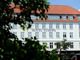På en hvid bygning med rødt tegltag og blå vinduer står der Hotel Europa på facaden, delvist indrammet af grønne grene.