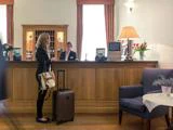 En kvinde med en kuffert står ved en hotelreception og taler med en receptionist. Lobbyen har blomster, en lampe og et hyggeligt siddeområde.