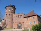 Et historisk murstensslot med et stort rundt tårn, buede trædøre og røde tegltage mod en klar blå himmel.