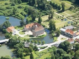Luftfoto af en stor herregård omgivet af en voldgrav, haver, træer og yderligere bygninger, med stier og grønne områder spredt ud over hele landskabet.