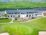 Et moderne hvidt golfklubhus med mørkt tag, omgivet af grønne plæner og sandbunkere, med parkerede biler og træer i baggrunden.