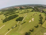 Luftfoto af en stor golfbane med grønne fairways, sandbunkers, træer og omkringliggende bygninger under en klar himmel.