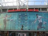 Forsiden af Manchester Uniteds stadion med store billeder af fodboldspillere, glasvinduer og et rødt Manchester United-skilt over indgangen.