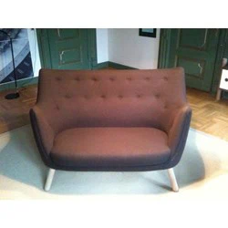 En moderne brun to-personers sofa med knappet ryglæn og lyse træben, placeret på et lyst tæppe i et rum med grønne døre og trægulve.