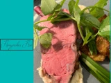 En tallerken med en tyk skive roastbeef, sprøde kartofler, grønne løg, bladgrønt og cremet puré på et gråt fad, ved siden af en blågrøn tekst, hvor der står Konversation Trio.