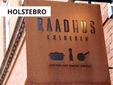 Et rustfarvet metalskilt med teksten RAADHUS KÆLDEREN med køkkenredskabsikoner og dansk tekst nedenunder er monteret på en murstensbygning. HOLSTEBRO står i en hvid boks over skiltet.