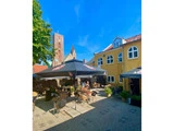 En gårdhave med udendørs café, borde, stole og store parasoller; en gul bygning og et kirketårn i baggrunden under en strålende blå himmel.