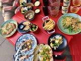 Et rødt bord med forskellige tallerkener med mad, herunder pommes frites, dumplings, stegt tun, grønne bønner, risretter, dypsaucer og drikkevarer, med to personer siddende.