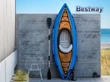 En blå oppustelig kajak med pagaj læner sig op ad en betonvæg i nærheden af en stålstol. Bestway-logoet ses i øverste højre hjørne. Havet og græsset er synligt i baggrunden.