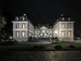 Et stort, elegant palæ med mange oplyste vinduer, set om natten. Bygningen har to fløje, symmetrisk arkitektur og en central indgang med en brostensbelagt sti foran.