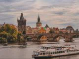 En turbåd sejler på en flod foran den historiske Karlsbro og tårnene i Prag med farverige bygninger og en overskyet himmel i baggrunden.
