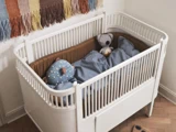 En hvid babyseng med en brun stødpude, blåt sengetøj og to tøjdyr ved siden af en væg med et farverigt vævet ophæng.