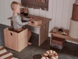 En ung dreng sidder ved et lille træskrivebord og leger med en æske og legetøj. Trælegetøj og stribede plysdyr ligger på gulvet og på en hylde ved siden af ham i et hyggeligt, neutralt farvet rum.