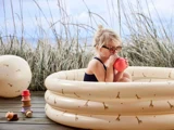 Et lille barn med solbriller sidder i en oppustelig pool med girafaftryk og holder en kop med græs og himmel i baggrunden.