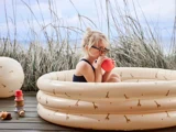 Et lille barn med briller sidder i en beige oppustelig pool med giraftryk og drikker af en kop, med højt græs og stablede legetøjskopper i nærheden.