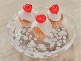 Tre små cupcakes med hvid glasur og et rødt kirsebær på toppen, arrangeret på en klar glastallerken med hvide polkaprikker, stillet på en lys træplade.