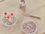 Et træbord med en klar glasplade med to små kager med kirsebær på toppen, en matchende glasskål, en træspatel og et glas chokolademælk.