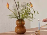En brun dobbeltvase med forskellige vilde blomster står på et træbord; en hånd tænder et blåt snoet lys i en guldlysestage i nærheden.