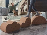 Et barn balancerer på legeklodser af kork formet som en sky, en spiral og en regnbue i et legerum med legetøj og et tæppe på gulvet.