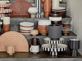 Et udvalg af moderne keramikservice i neutrale og jordfarvede toner arrangeret på betontrin, herunder tallerkener, krus, skåle og stribede stativer.