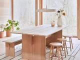 Et spisebord i lyst træ med taburetter og en bænk står ved store vinduer. Enkle vaser med blomster og potteplanter pynter i det minimalistiske, luftige rum.