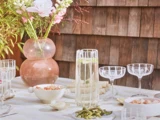 Et solbeskinnet bord udendørs, dækket med glas, en kande vand, skåle med mad og en lyserød vase med blomster, med en væg af træspån i baggrunden.