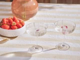 To coupéglas med klare drikkevarer og isbiter med blomstermønster på en stribet dug ved siden af en skål med jordbær og en glasvase i sollyset.