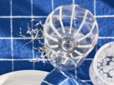 Et klart glas med hvide striber og tørrede blomster indeni står på en blå ternet dug ved siden af et andet mønstret glas og en hvid tallerken.