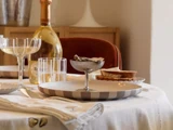 Et bord dækket med en stribet tallerken, et glas med østers, vinglas, en flaske mousserende vin og brødskiver på en hvid dug.