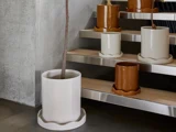 Flere store keramiske blomsterkrukker i hvidt og brunt står på trætrapper og på gulvet, hvoraf nogle indeholder tynde trægrene, i et moderne, minimalistisk interiør.