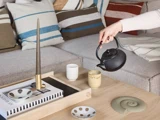 En person skænker te fra en sort tekande over i en keramisk krus, der står på et træsofabord med bøger, et stearinlys og pyntegenstande, foran en sofa med stribede puder.