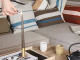 En hånd tænder et højt stearinlys på et sofabord, hvor der står kopper, et blad og en tekande. Bagved står en beige sofa med stribede puder.