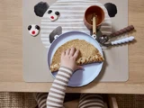 Et barn i stribet pyjamas rækker ud efter en sammenfoldet pandekage på en tallerken, mens der på bordet ligger en bordskåner med pandamotiv, en kop, bestik og kager formet som pandahoveder.