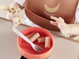 Et barn, der har en hagesmæk med bananmotiv på, har rigatoni-pasta på fingrene og rækker ud mod en skål med mere pasta og en gaffel.