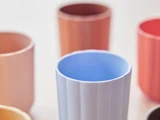 Fem plastikkrus i pastelfarver, herunder blå, orange, lyserød og rød, er anbragt på en lys overflade. Det blå krus er i fokus midt i billedet.